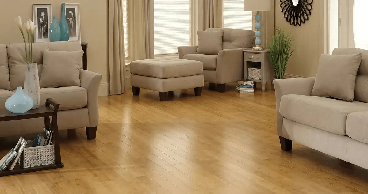 A living room with professionally installed bamboo flooring, beige sofas, a matching ottoman, a side table with a blue lamp, vases, framed pictures, and large windows with blinds and curtains in Pittsburgh, PA